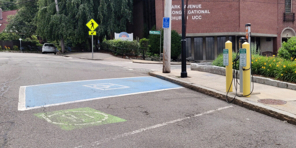 An electric vehicle charging station, near the Park Ave Congregational Church.  The spaces is marked with faded green and white paint. The vehicle charger is located behind two yellow bollards.