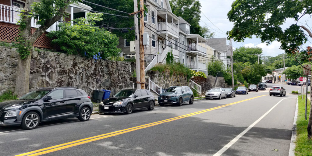 A street where the houses are approximately 10 feet above the sidewalk level. There are retaining walls next to the sidewalk, and stairways leading up to the houses.  A row of parked cars sits alongside the curb.