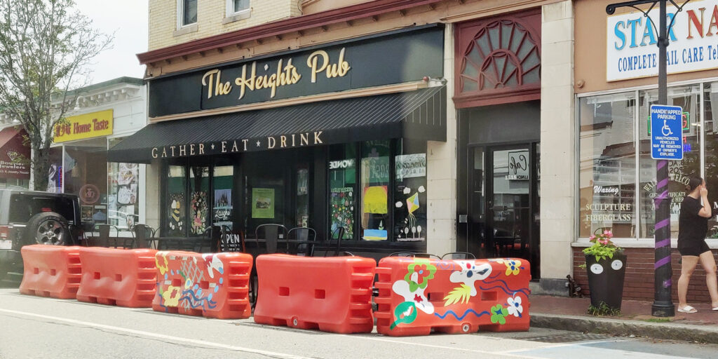 A curbside dining space that's surrounded by orange traffic barriers.  The restaurant has a sign with the words "The Heights Pub", and a black awning with the words "Gather, Eat, Drink".