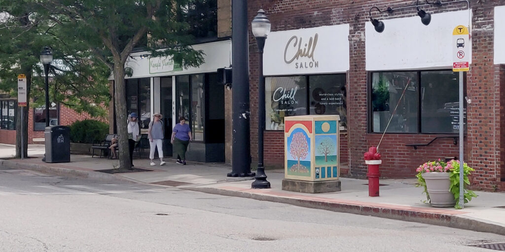 A curb-side bus stop.  The bus stop is marked with a sign. There are trees, planters, and storefronts in the photo.