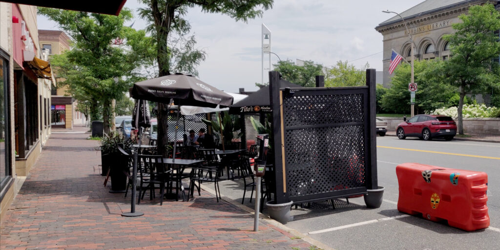 A pair of curbside parking spaces that have been converted to an outdoor dining area.  There are tables, chairs, umbrellas, and a wooden screen.